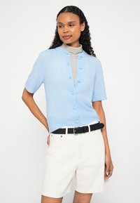 Gaiši zils īss piedurkņu kardigans ar lielām pogām, apaļu apkakli, valkāts virs svītrainas augstas apkakles džempera, kombinēts ar baltām šortiem un melnu jostu.