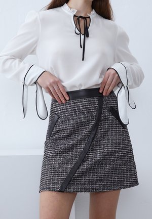 Vrouw draagt witte blouse met zwarte dasdetail en uitlopende manchetten, gecombineerd met zwart-witte getextureerde minirok met namaakleren rand.