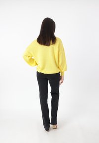 Un maglione a maglia giallo oversize con maniche lunghe, abbinato a pantaloni neri attillati e tacchi, su uno sfondo bianco semplice.