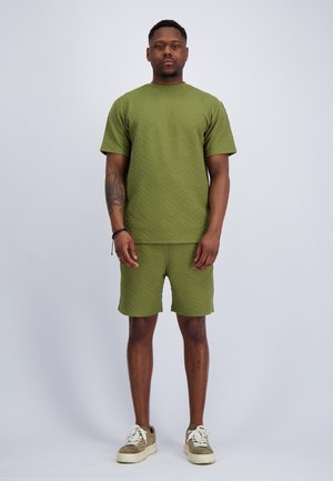 Mann steht mit Blick nach vorne, trägt eine strukturierte olivgrüne Kurzarmshirt und passende Shorts mit beigen Sneakern vor einfarbigem Hintergrund.