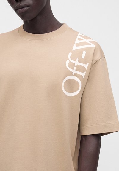 Personne à la peau foncée portant un t-shirt beige oversize avec "Off-White" imprimé en blanc sur l'épaule gauche et la poitrine.