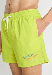 Limetkově zelené plavky s elastickým pasem, bílou šňůrkou na stažení a vyšitým textem na dolní straně, který obsahuje barevné písmo.