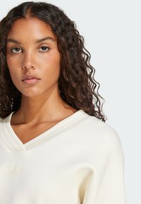 Vit, texturerad V-ringad sweatshirt med tre horisontella ränder ovanför halsringningen och en diskret präglad logotyp på bröstet.