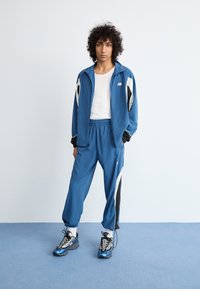 En person med krøllete hår står på en blå gulv iført en blå og hvit treningsdress med hvit skjorte og matchende blå joggesko.