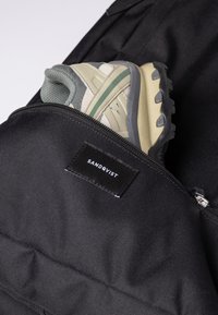 Black backpack featuring a zipper closure, with a light-colored sneaker partially visible in a side compartment. Brand label "SANDQVIST" displayed.