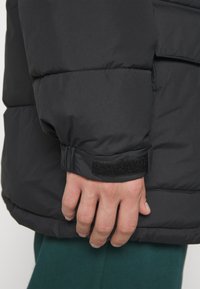 Mano descansando al lado, vistiendo una chaqueta acolchada negra con puños de Velcro ajustables y pantalones verdes visibles en la parte inferior.