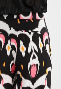 Gros plan sur un pantalon avec un motif ikat abstrait en noir, blanc, rose et orange et une poche latérale, porté avec un haut noir transparent.