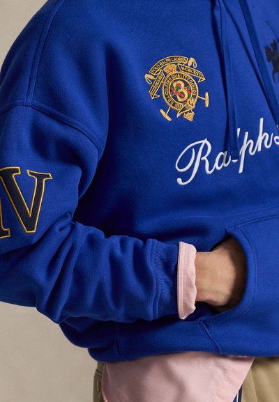 Personne portant un sweat à capuche Polo Ralph Lauren bleu avec un blason brodé et du texte, une main dans la poche avant, le poignet d'une chemise rose visible.