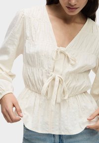 Blusa bianca strutturata con scollatura a V profonda, vita arricciata e chiusura con fiocco. Maniche lunghe con polsini elasticizzati. Vestibilità casual.