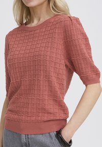 Frau trägt ein rostfarbenes, kurzärmeliges Strickoberteil mit strukturiertem Muster, kombiniert mit grauer Hose und einem schwarzen Armband am Handgelenk.