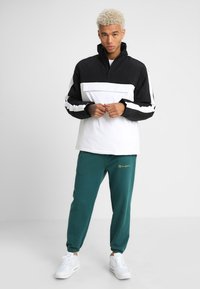 Svart og hvit half-zip jakke med frontlomme, sammen med teal joggebukser med gullbroderi, komplementert av hvite sneakers.