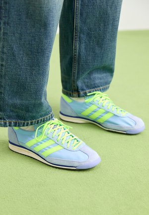 Šviesiai mėlyni sportiniai bateliai su žaliais akcentais, pagaminti iš zomšos ir sintetinės medžiagos, su trimis juostelėmis ir tekstūruotu padu. Derinami su laisvais džinsais.