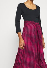 Svart långärmad topp kombinerad med en rosa och mörkblå mönstrad kjol med en knutdetalj i midjan och en utsvängd silhuett.