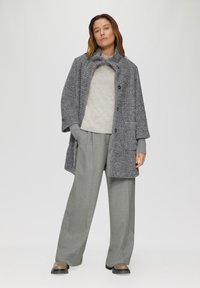 Manteau gris texturé avec de grands boutons avant, large col et poches latérales ; associé à un pull gris clair et un pantalon gris ample.