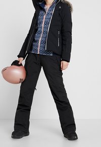 Osoba w czarnych spodniach narciarskich, czarnej kurtce z futrzanym kapturem, niebieskiej bluzie zapinanej na zamek z wzorem, trzyma różowy kask narciarski z boku.