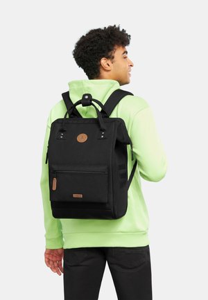Schwarzer Rucksack mit rechteckiger Form, strukturiertem Stoff, vorderer Reißverschlusstasche, Lederelementen und verstellbaren Trägern. Wird von einer Person in einem grünen Hoodie getragen.
