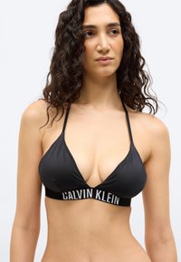 Γυναίκα με σγουρά σκούρα μαλλιά που φοράει μαύρο μαγιό halter Calvin Klein, κοιτώντας ελαφρώς προς το πλάι έναντι απλού φόντου.