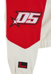 Manche de veste rouge et blanche avec un logo "05" brodé et un petit patch "L" noir et rouge près du poignet.