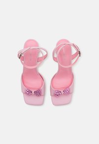 Sandales à plateforme roses avec des brides croisées, dotées d'une finition brillante, d'une semelle plate et d'un nœud décoratif orné de strass à l'avant.