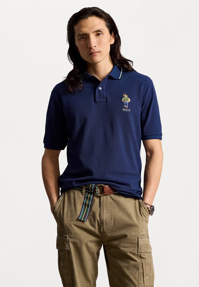 Granatowa koszulka polo z krótkimi rękawami i prążkowanym kołnierzykiem, ozdobiona zielonymi i białymi akcentami, haftowana kolorowym logo na klatce piersiowej.