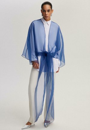 Vrouw die een wit shirt en broek draagt met een doorschijnende donkerblauwe sjaal gestrikt rond de taille, staand tegen een effen lichte achtergrond.