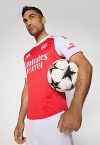 Rode en witte korte mouw voetbalshirt met een textuurdesign, voorzien van het logo "Emirates Fly Better". Houdt een zwart-wit voetbalsbal met sterrenpatroon vast.