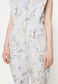 Floral jumpsuit featuring a light gray fabric with blue and green botanical prints, elastic waistband, and wide shoulder design.