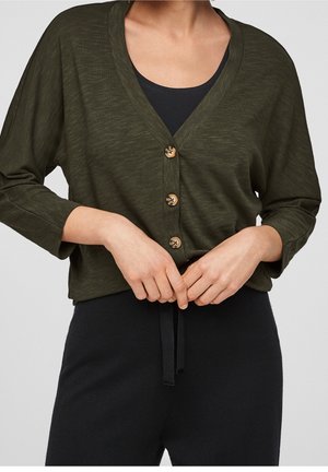 Osoba ubrana w ciemnooliwkowy kardigan na guziki, nosząca czarny top, z obiema rękami złączonymi przed sobą na wysokości talii.