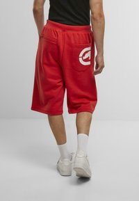 Red cotton shorts with an elastic waistband and side pockets, featuring a large white logo on the back pocket. Worn with white sneakers and socks.