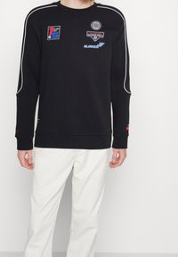 Sudadera negra con ribetes blancos, que presenta múltiples parches bordados. Tiene mangas largas y un cuello y puños de canalé.