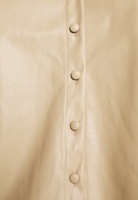 Beige läderplagg med knappstängning, med en jämn textur och subtil lyster. Knapparna är rundade och matchar färgen på tyget.