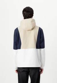 Waterdichte jas met een kleurblokkenschema: beige, marineblauw en wit. Voorzien van een capuchon en elastische manchetten, gemaakt van lichtgewicht materiaal.