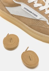 Beige semsket sko med hvite skinnaksenter, perforert design og gummisåle. Inkluderer to rullede beige skolisser.