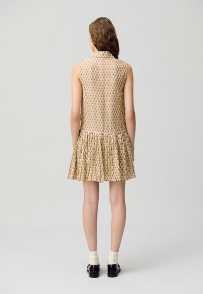Vestido sin mangas beige con un patrón floral, falda recogida y detalle en el cuello. Usado con calcetines blancos y zapatos negros.
