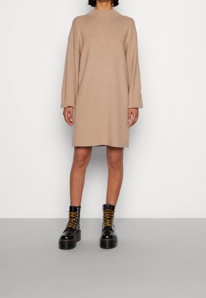 Abito a maglia beige con maniche lunghe, colletto alto e una vestibilità rilassata, abbinato a stivali da combattimento neri con lacci e dettagli gialli.