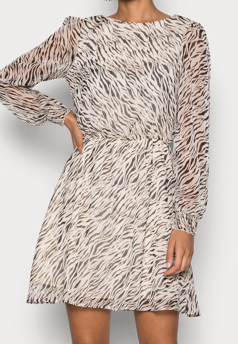 Femme portant une mini-robe à manches longues à imprimé zèbre beige et marron, taille cintrée et manches transparentes, posant avec une main sur la hanche.