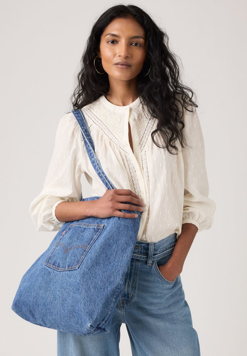 Γυναίκα με μακριά σγουρά μαλλιά που φοράει λευκό πουκάμισο και τζιν, κρατώντας μια μεγάλη μπλε τσάντα ώμου από τζιν με πίσω τσέπη.