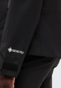 Μανίκι μαύρου μπουφάν με λευκό λογότυπο GORE-TEX κοντά στη ρυθμιζόμενη μάνικα με Velcro στο χέρι ενός ατόμου, τονίζοντας τη σήμανση αδιάβροχου υφάσματος.