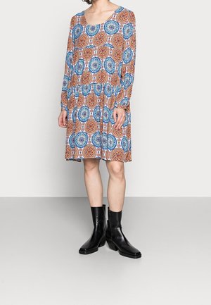 Kobieta ubrana w sukienkę z długim rękawem do kolan w niebiesko-pomarańczowe kołowe wzory, stojąca na szarej podłodze w czarnych butach na obcasie do połowy łydki.