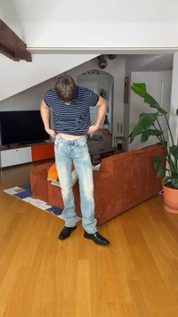 Νεαρός άνδρας με ριγέ πουκάμισο και τζιν ρυθμίζει το παντελόνι του σε ένα μοντέρνο σαλόνι με ξύλινο πάτωμα, πορτοκαλί καναπέ και μεγάλο γλαστρόφυτο.