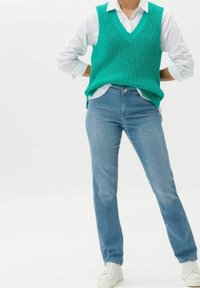 Turkos stickad ärmlös tröja med V-ringning över en vit skjorta, kombinerad med ljusblå raka jeans och vita sneakers.