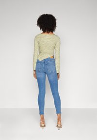 Parte superior de manga larga texturizada verde con detalle de espalda retorcida, combinada con jeans ajustados azules y sandalias de tacón color beige. No se ven la parte frontal ni los lados.