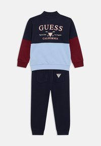 Διπλό σετ φόρμας σε μπλε ναυτικό και βυσσινί χρώμα με ανοιχτό γαλάζιο πάνελ· διαθέτει ψηλό γιακά, ριμπ μανσέτες και κεντημένο λογότυπο στο πάνω μέρος.