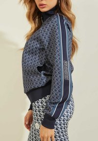 Giacca blu navy con un design a motivo e righe in contrasto, con colletto alto e polsini a banda, che presenta il marchio "GUESS".