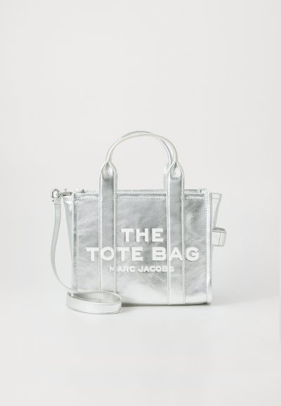 Sac à main en métal argenté avec deux poignées supérieures, une bandoulière détachable et un texte de logo blanc audacieux à l'avant. Texture lisse, forme rectangulaire.