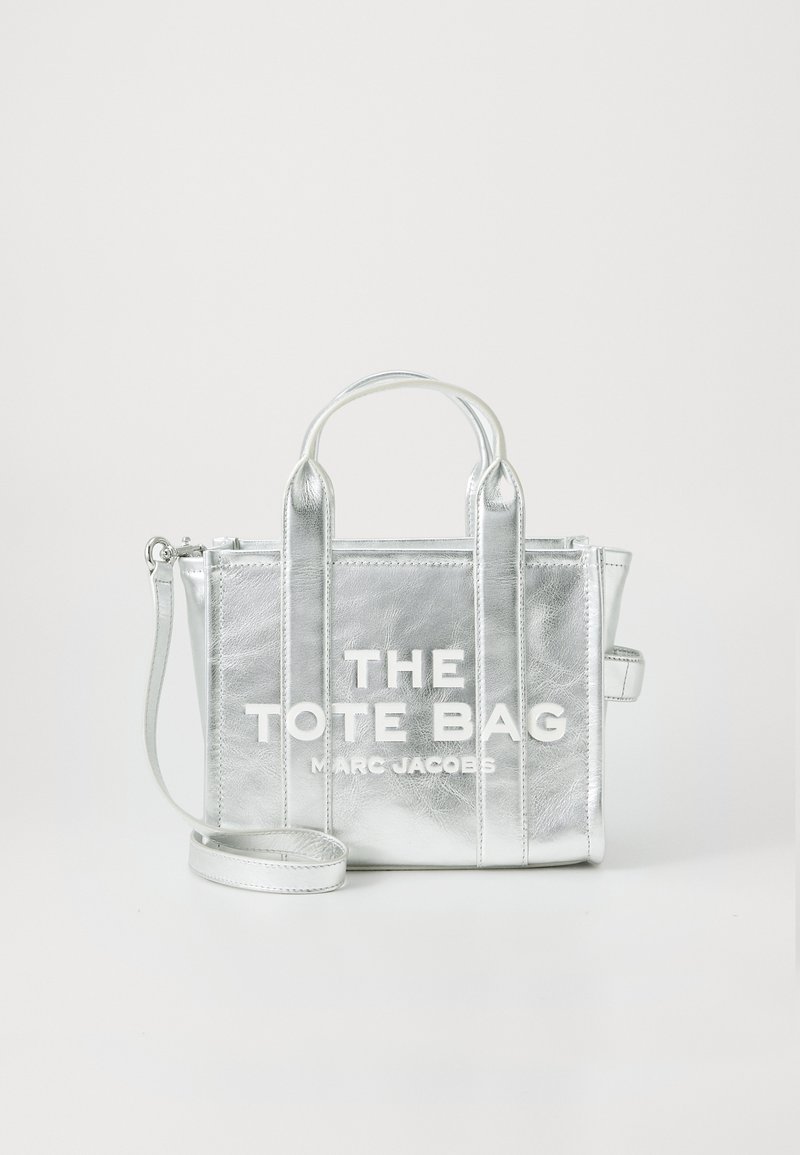Saco tote metálico prateado com duas alças superiores, alça de ombro destacável e texto em branco em destaque na frente. Textura suave, forma retangular.