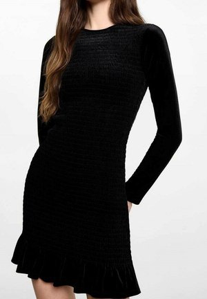Kobieta w dopasowanej, czarnej sukience z długim rękawem o teksturowanym wzorze i falbaniastym dołem, stojąca na tle jednolitego białego tła.