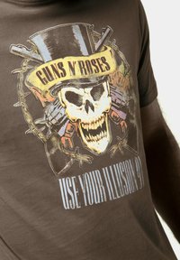 Brun bomulds t-shirt med et grafisk motiv af et kranium i en høj hat med våben, omgivet af dekorative elementer og teksten "GUNS N' ROSES."