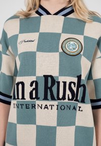 Nærbillede af grøn og beige ternet skjorte med stribet krave og ærmekanter, tekst "In a Rush INTERNATIONAL" og rund emblemmærkat.