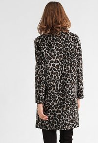 Mujer con cabello hasta los hombros que lleva un abrigo de leopardo de longitud a la rodilla, de pie de espaldas contra un fondo liso.